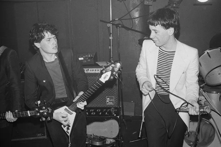 Black and white still of two young white men on a small stage singing and playing the guitar in the 80s