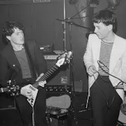 Black and white still of two young white men on a small stage singing and playing the guitar in the 80s