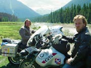 Two white men with scraggly facial hair sit on their motorbikes in a dramatic green mountainous and forest landscape