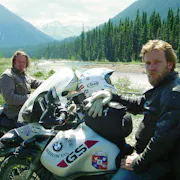 Two white men with scraggly facial hair sit on their motorbikes in a dramatic green mountainous and forest landscape