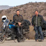 Two white men in thick clothing standing beside their motorcyclists in a dry shrub landscape