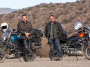 Two white men in thick clothing standing beside their motorcyclists in a dry shrub landscape