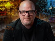 A middle aged bald white man with black framed glasses looks down the camera as neon multi-coloured lines and text swirl around him
