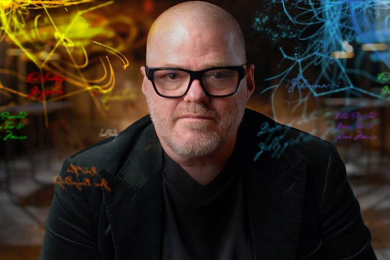 A middle aged bald white man with black framed glasses looks down the camera as neon multi-coloured lines and text swirl around him