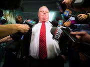 Large white man in suit with bright red tie with a many handheld microphones in front of him.