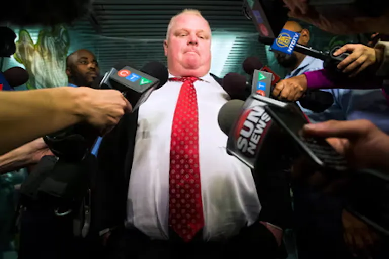 Large white man in suit with bright red tie with a many handheld microphones in front of him.