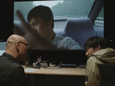 Two men in conversation with one another sit at a table with their backs to us. One of the men at the table is wearing glasses and a black shirt, and the other is wearing a grey-brown raincoat. The table has audio-visual equipment on it. Above the table is a large screen with the image of a man sitting in the back of a car.