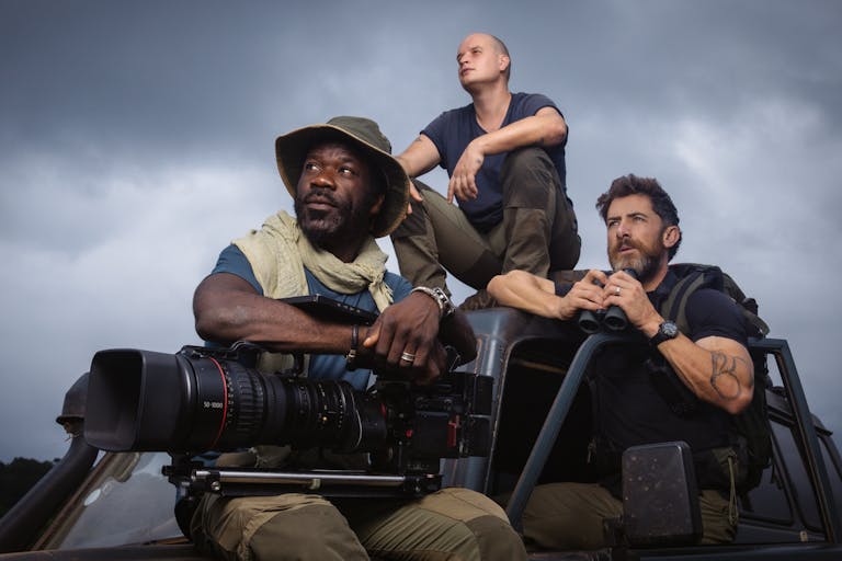 A Black man and two white men sit on an offroad jeep in explorer clothing holding a large camera