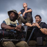 A Black man and two white men sit on an offroad jeep in explorer clothing holding a large camera