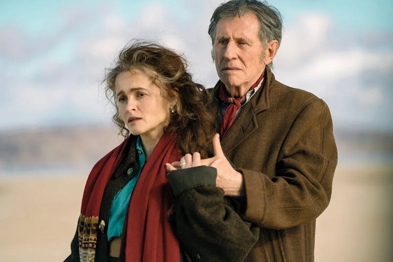 A middle aged white woman and man in coats and scarves standing in a light embrace on a beach