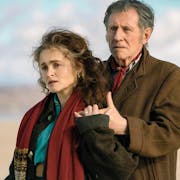 A middle aged white woman and man in coats and scarves standing in a light embrace on a beach