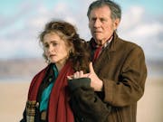 A middle aged white woman and man in coats and scarves standing in a light embrace on a beach