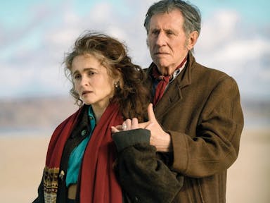 A middle aged white woman and man in coats and scarves standing in a light embrace on a beach