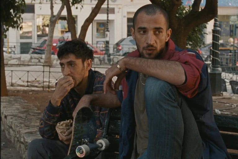 Two young Palestinian men sit under a tree with a skateboard propped up next to them looking forlorn