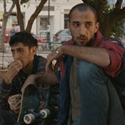 Two young Palestinian men sit under a tree with a skateboard propped up next to them looking forlorn