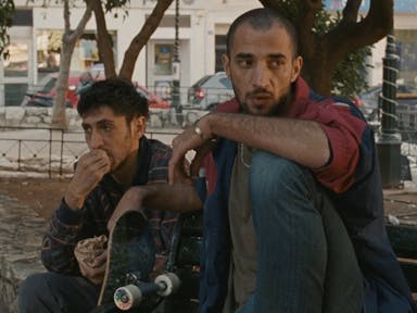 Two young Palestinian men sit under a tree with a skateboard propped up next to them looking forlorn