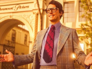 A white man with thick rimmed glasses, check suit and a wide red tie smiling to someone at camera left