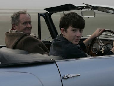 Two white men, a younger and an older one, driving a convertible blue car looking at the camera