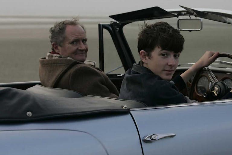 Two white men, a younger and an older one, driving a convertible blue car looking at the camera