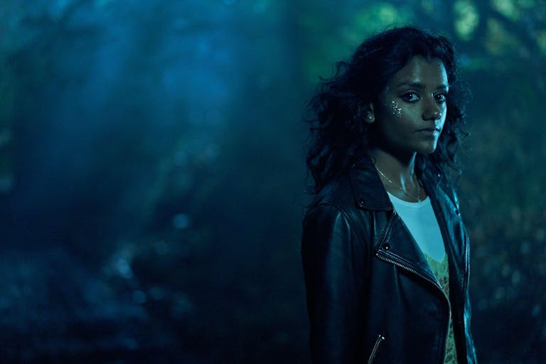 A woman in leather jacket stands in a misty forest in the dark looking at the camera