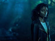A woman in leather jacket stands in a misty forest in the dark looking at the camera