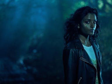 A woman in leather jacket stands in a misty forest in the dark looking at the camera