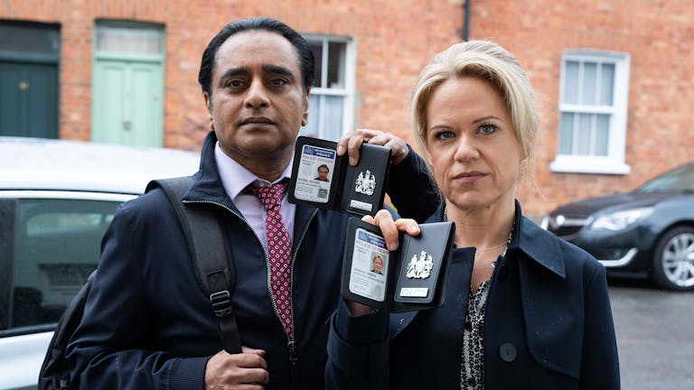 A middle aged brown man and middle aged white woman standing outside a house, showing metropolitan police badges, looking serious