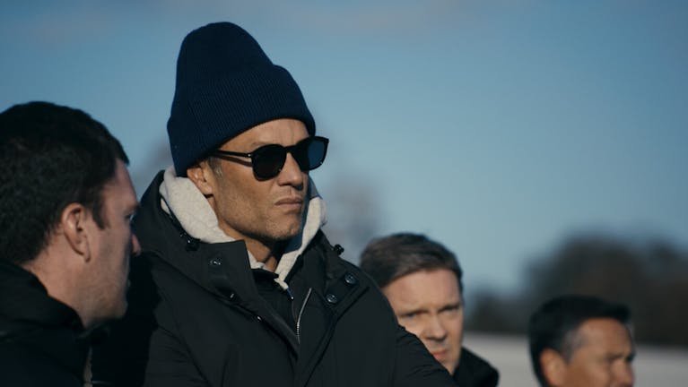 A tanned white man in a thick coat, sunglasses and a tall beanie hat talking to other white men