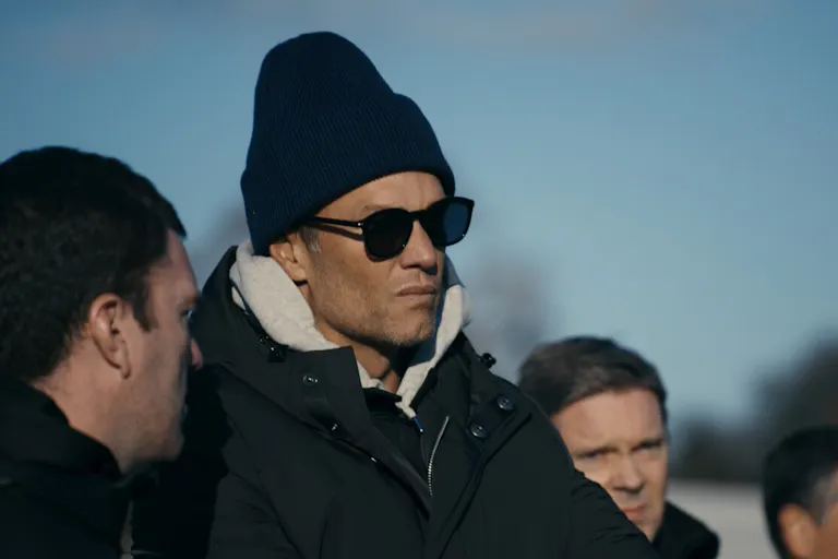 A tanned white man in a thick coat, sunglasses and a tall beanie hat talking to other white men
