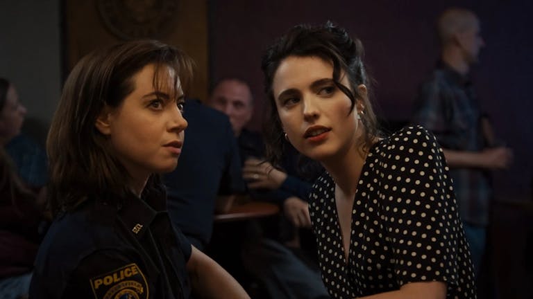 Two young women in a dark bar, one in a police uniform the other in a black polka dot dress looking quizzically over their shoulders 