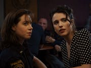 Two young women in a dark bar, one in a police uniform the other in a black polka dot dress looking quizzically over their shoulders