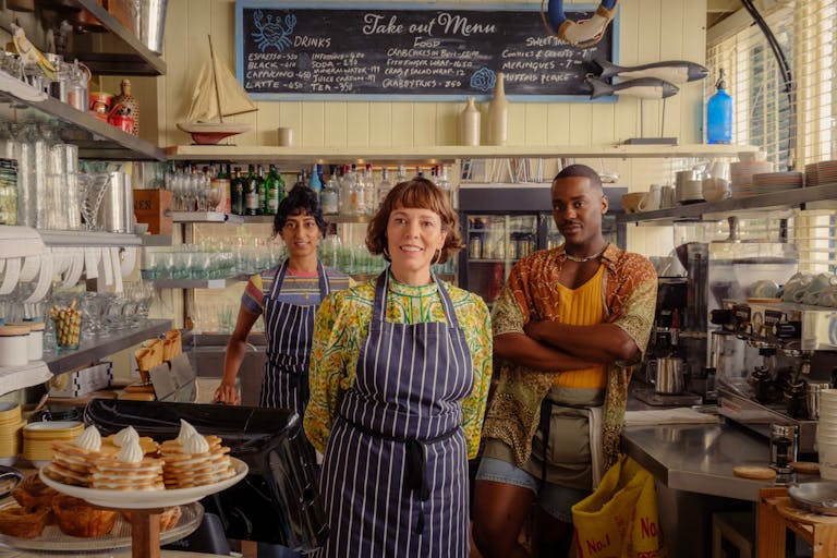 A middle aged white woman, a young Black man and a young brown woman stand in stripey aprons behind the counter of a café