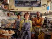 A middle aged white woman, a young Black man and a young brown woman stand in stripey aprons behind the counter of a café