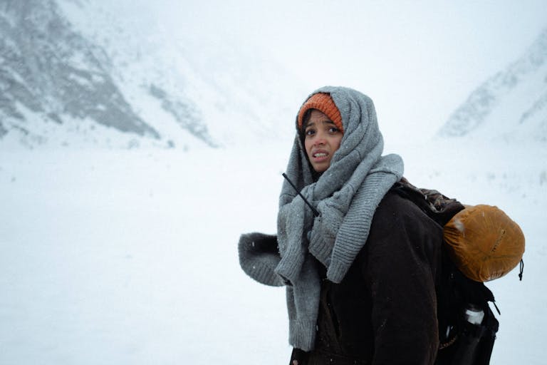 A young Black woman in a blizzard, snowy landscape wearing layers of woollen clothing, carrying a rucksack and sleeping bag, looking agitated