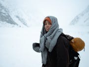 A young Black woman in a blizzard, snowy landscape wearing layers of woollen clothing, carrying a rucksack and sleeping bag, looking agitated