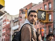 A British Pakistani man in a tan jacket crossing the street in New York looking over his shoulder
