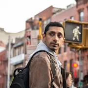 A British Pakistani man in a tan jacket crossing the street in New York looking over his shoulder
