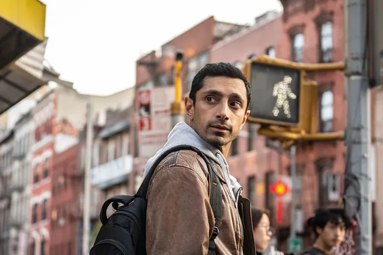 A British Pakistani man in a tan jacket crossing the street in New York looking over his shoulder