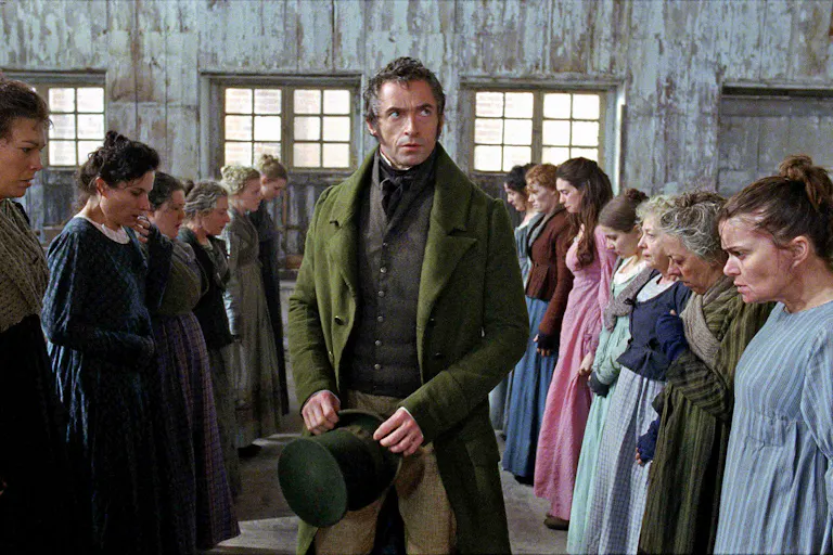 A white man in smart green velvet suit and top hat walks between two roles of woman in simple dresses standing looking nervous on a factory floor