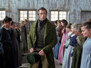 A white man in smart green velvet suit and top hat walks between two roles of woman in simple dresses standing looking nervous on a factory floor