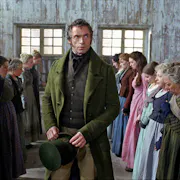 A white man in smart green velvet suit and top hat walks between two roles of woman in simple dresses standing looking nervous on a factory floor