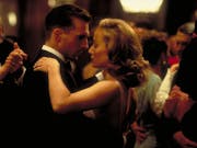 A white man in a suit dances closely with a white woman in a black dress as they look at each other longingly