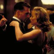 A white man in a suit dances closely with a white woman in a black dress as they look at each other longingly