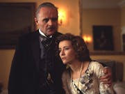 A middle aged white man with his arm around a younger white woman comforting her in a grand manor house, wearing Edwardian clothes