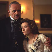 A middle aged white man with his arm around a younger white woman comforting her in a grand manor house, wearing Edwardian clothes