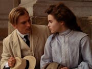 A young white man in a sand coloured suit and hat and a young white woman in a high necked light blue dress sit on stone steps looking at each other