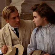 A young white man in a sand coloured suit and hat and a young white woman in a high necked light blue dress sit on stone steps looking at each other