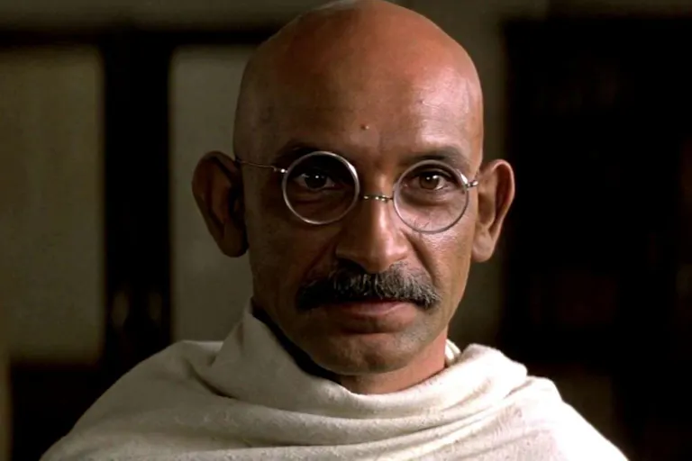A bald brown man with round glasses and a small moustache wrapped in a white cloth