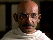 A bald brown man with round glasses and a small moustache wrapped in a white cloth