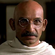 A bald brown man with round glasses and a small moustache wrapped in a white cloth
